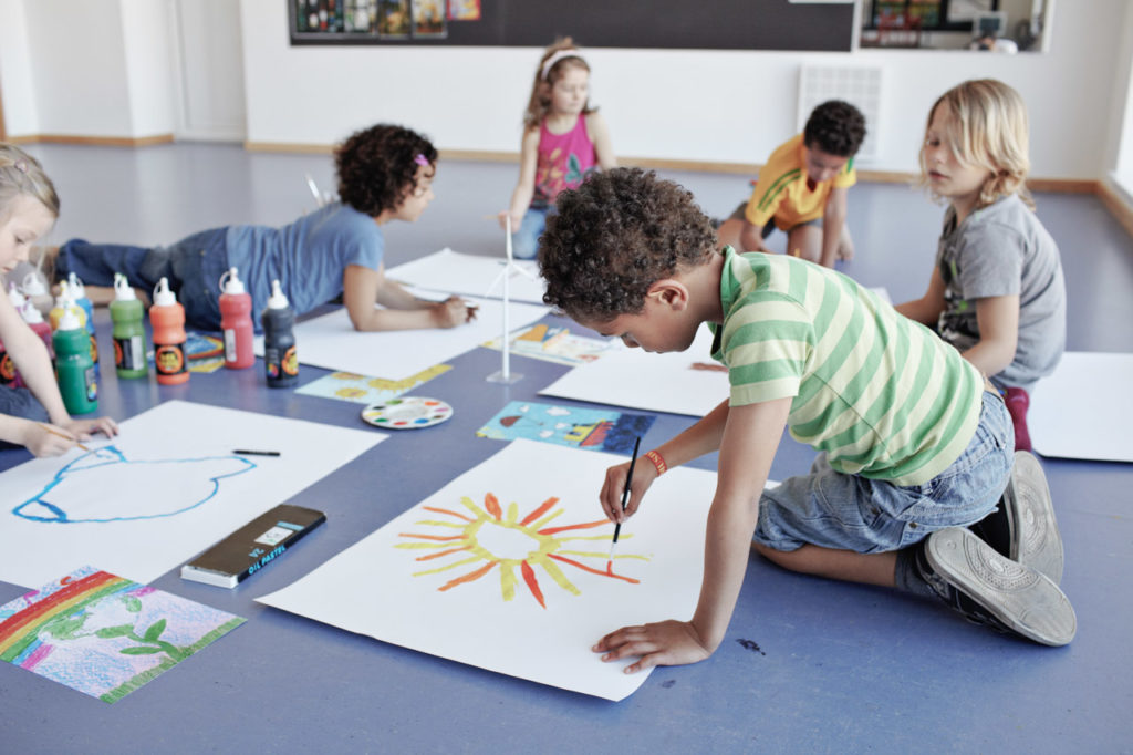 Young children sit on the floor painting with water colors on large white poster boards during a community event