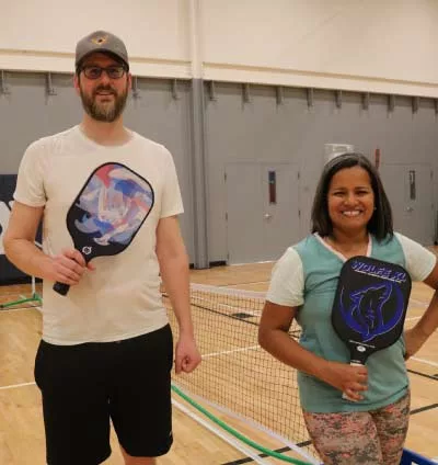 A tall man in a white shirt and a much shorter woman in a blue fitness top smile while holding pickleball paddles on the courts at YMCA of the North Shore