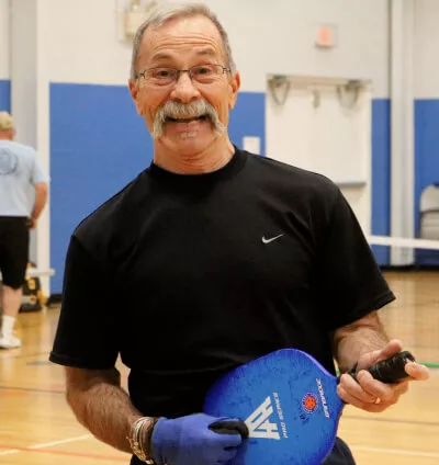 A YMCA community member in a black Nike shirt goofs off holding a pickleball racquet and strumming at it as if it were an electric guitar