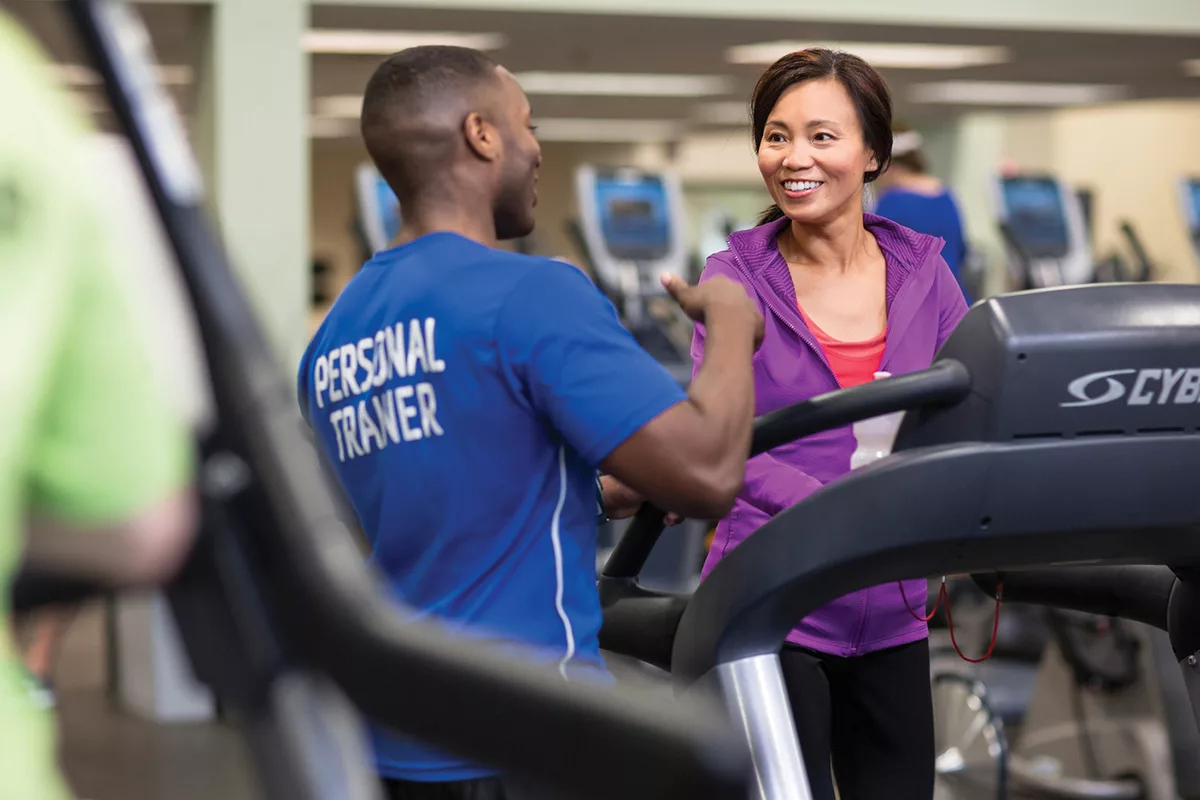A personal trainer at YMCA North Shore speaks to a smiling woman on a treadmill during their workout coaching session