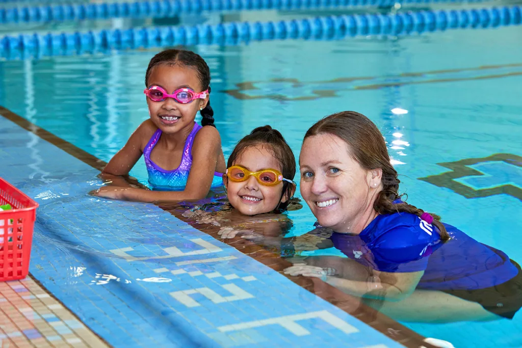Two small girls and a woman smile while sitting in the pool using their hands on the wall to support themselves during swim lessons
