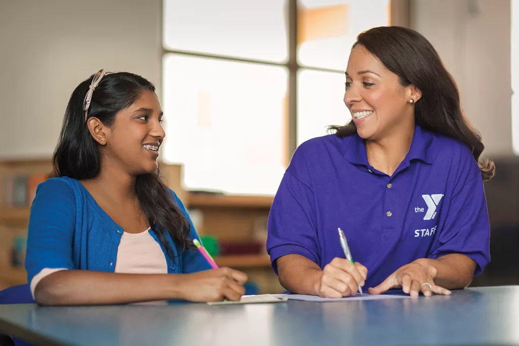 A young woman hold a pencil while a YMCA leader in a purple polo smiles and demonstrates during educational instruction