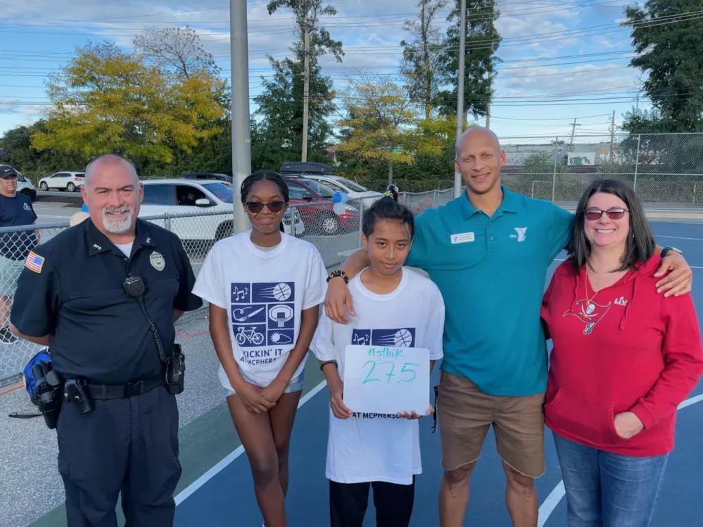 YMCA Teen Program McPherson Asthik holds a sign reading '27.5' as a YMCA leader puts his arm around he and another adult women wearing a red Buccaneers hoodie. Another young woman and a police office stand to his right.