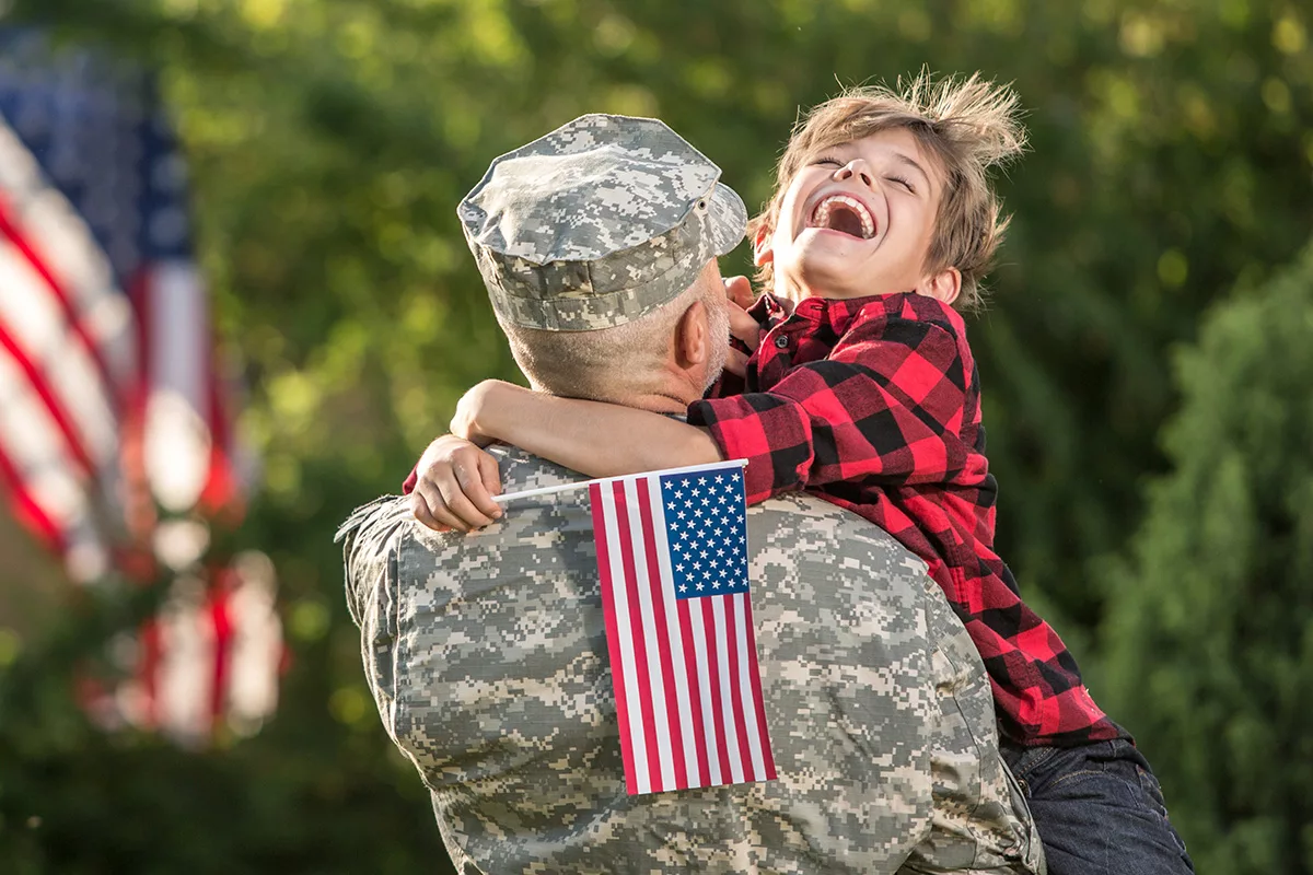 A young boy in a red flannel shirt laughs with joy as he hugs his father, a soldier in uniform, while holding an American flag