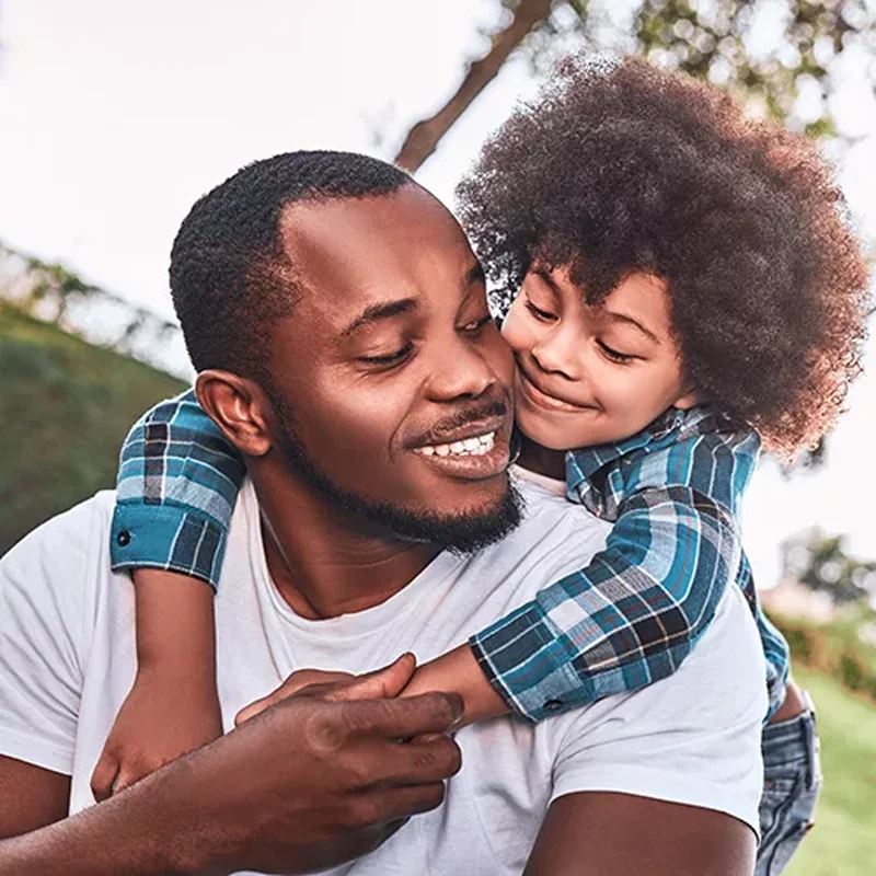 A young child holds onto his father's neck as the father looks back smiling and holding the son's arm for support