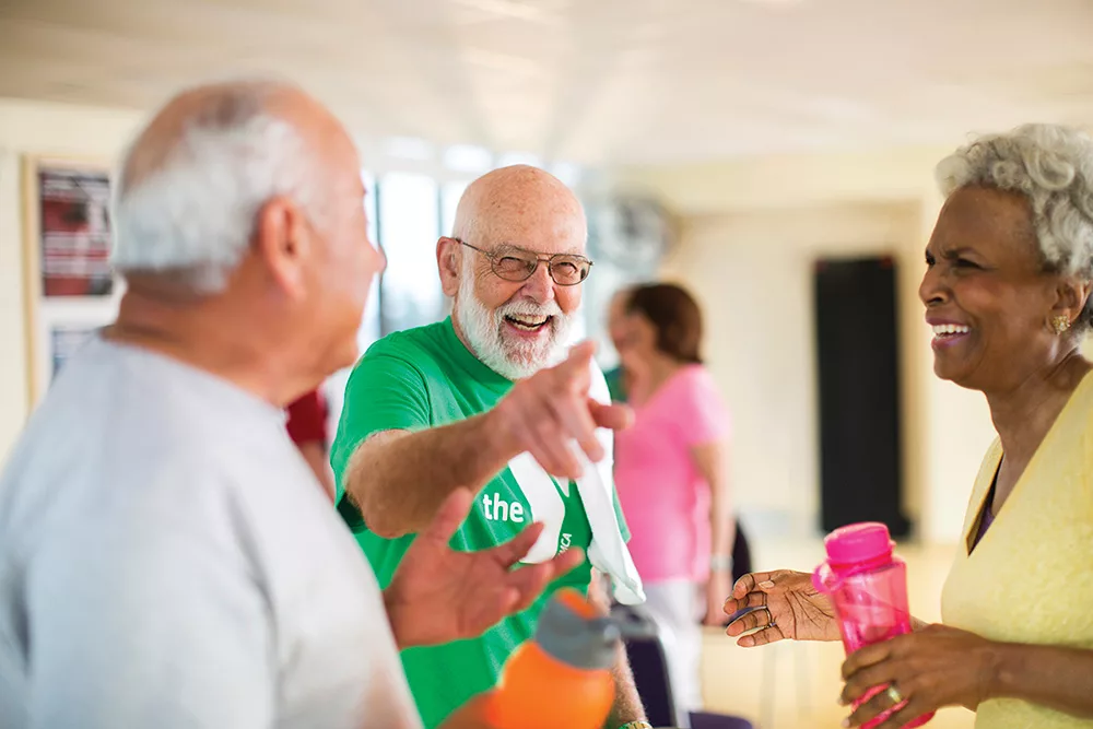 A senior man in a green YMCA shirt smiles while laughing and pointing at the camera as a woman in a yellow shirt and another man talk in the foreground.