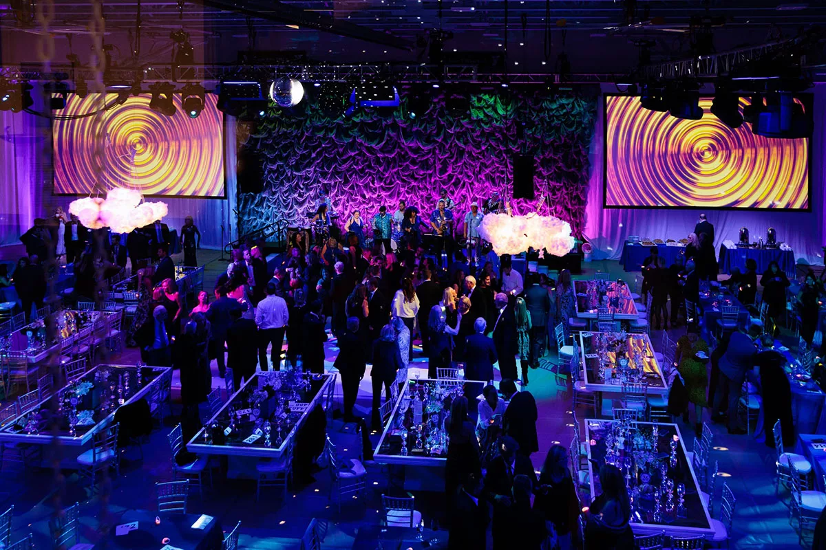 A overhead vantage point of guests enjoying the dance floor at the YMCA of the North Shore Wanderlust Ball while a brand in blue performs and large screens on either side of the stage show visuals to match the tempo.