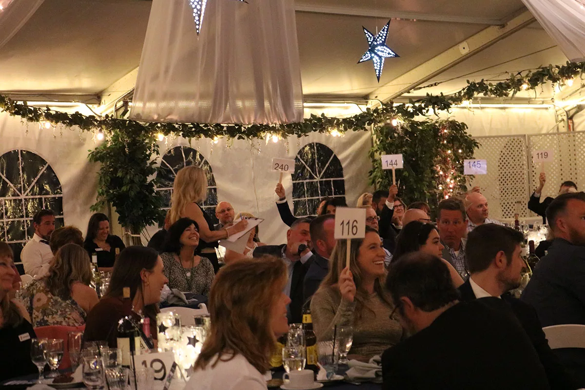 Guests of the YMCA Legacy Gala hold up bidding numbers while seated at formal tables in an event-hall style tent.