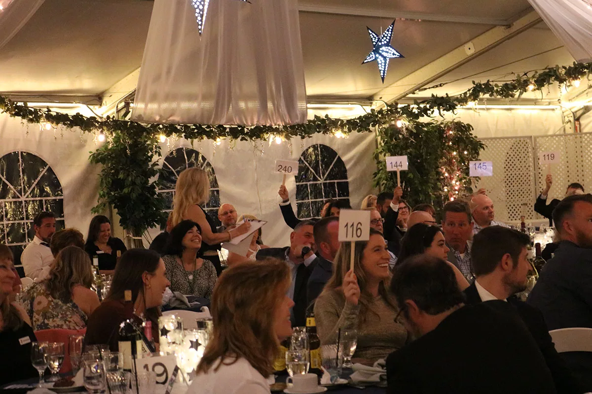Haverhill YMCA Legacy Gala YMCA of the North Shore Guests of the YMCA Legacy Gala hold up bidding numbers while seated at formal tables in an event-hall style tent.