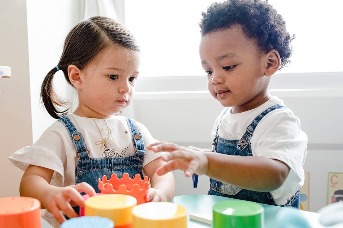 Two toddlers move large, colorful blocks around wearing matching white shirts and overalls at childcare.